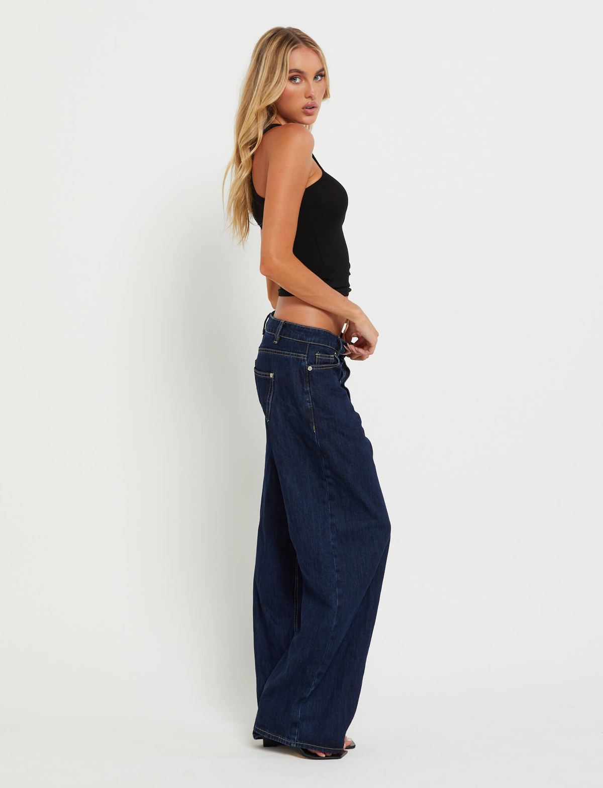 A woman with long blonde hair stands sideways against a white background, wearing TIGERMIST FELIX JEAN - DARK DENIM with an oversized fit, a black tank top, and high heels, looking toward the camera.