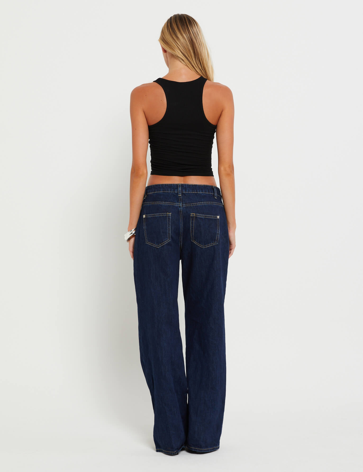 A woman with long hair stands against a white background, wearing the TIGERMIST FELIX JEAN - DARK DENIM in an oversized fit and a black sleeveless crop top, facing away from the camera.