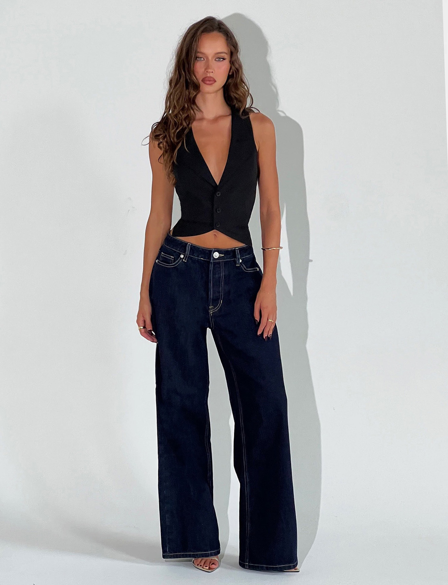 A woman with long wavy hair and a neutral expression wears TIGERMIST’s FELIX JEAN - DARK DENIM, a high-waisted, wide-leg stretch denim, paired with a sleeveless black buttoned vest against a plain white background.