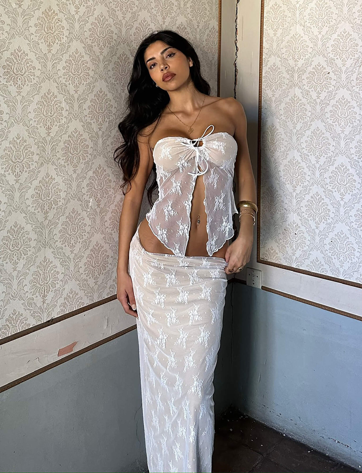 A woman with long dark hair poses in an ornate room with patterned wallpaper and wainscoting. She is wearing a TIGERMIST strapless white lace top tied in the front and the KAROLINA SKIRT - WHITE: WHITE LACE. She accessorizes with gold bangles and stands with one foot slightly forward.