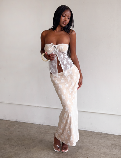 A woman poses indoors wearing the TIGERMIST KAROLINA TOP in white lace, a semi-sheer strapless top with a center bow and delicate floral pattern, paired with a matching long skirt. She accessorizes with white heels and a chunky bracelet.