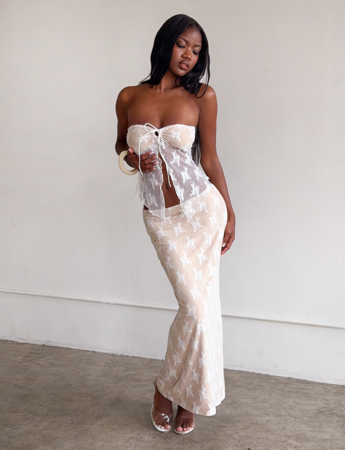 A woman models the TIGERMIST KAROLINA SKIRT - WHITE in white lace, paired with a matching sheer white lace top and white heels, posing indoors against a plain wall.