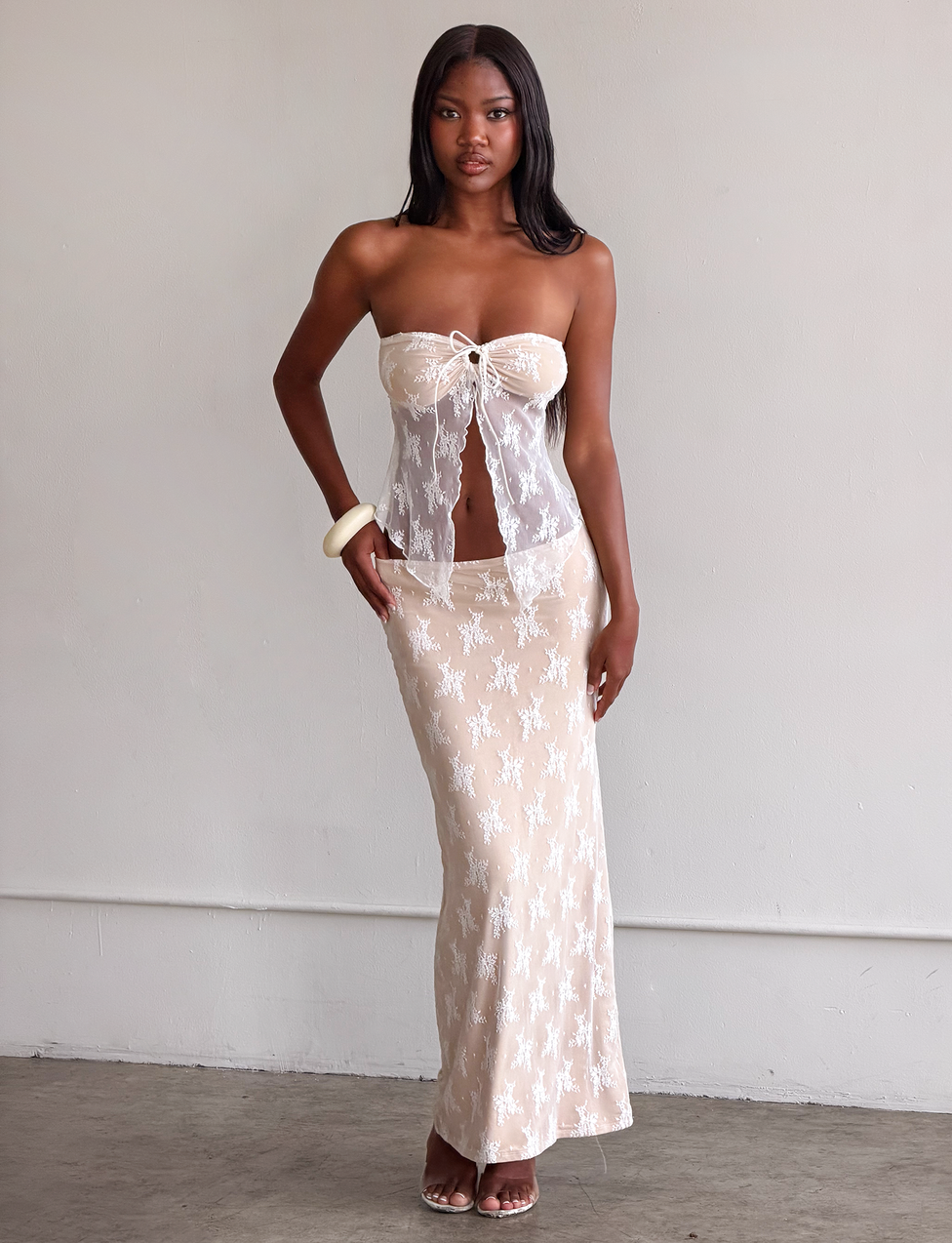 A woman stands indoors against a plain wall, wearing the TIGERMIST KAROLINA SKIRT - WHITE: WHITE LACE with a matching sheer lace strapless top. She has long straight hair, a white bangle, and open-toe heels.