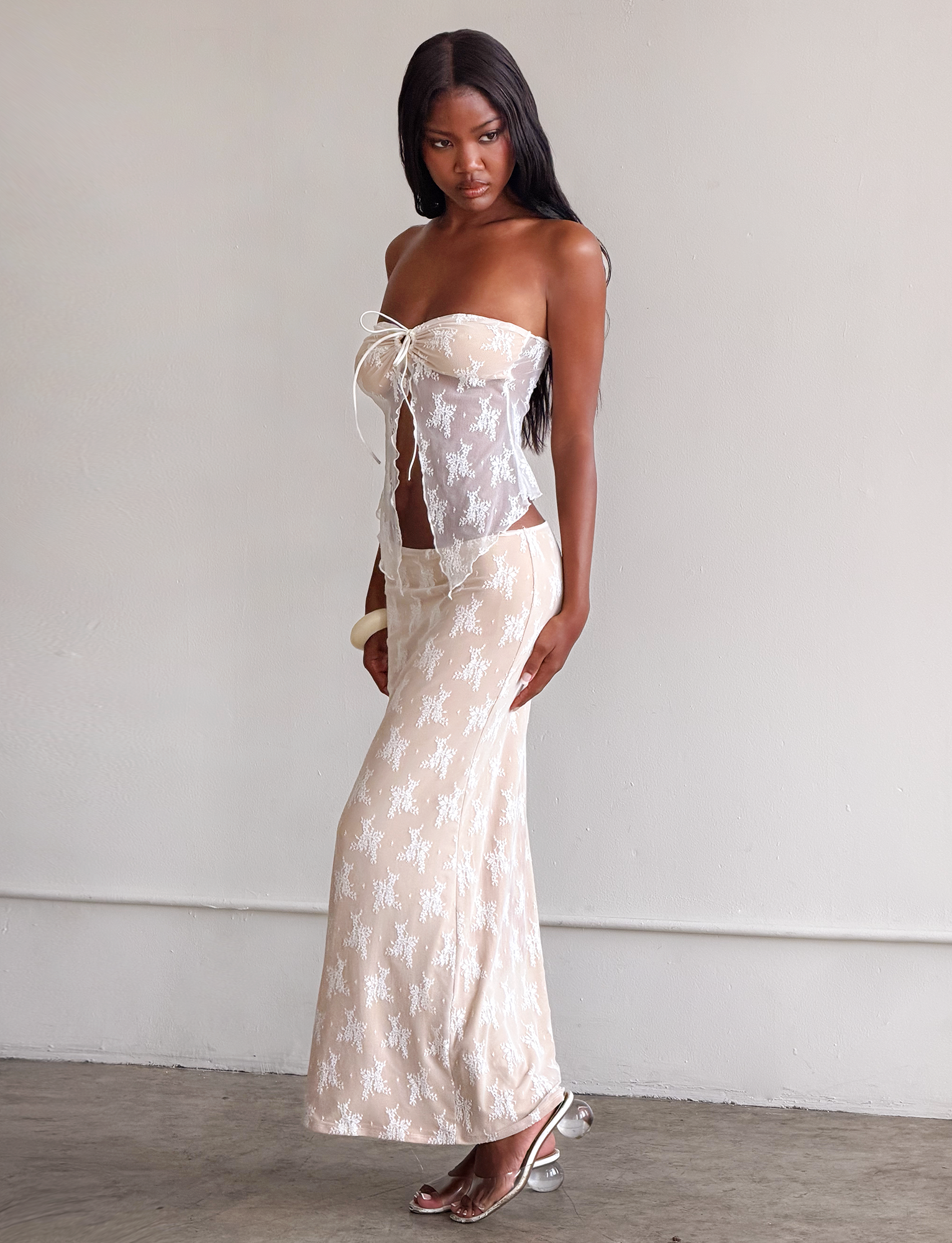 A woman with long dark hair wears the TIGERMIST KAROLINA TOP - WHITE, a strapless white lace top with a front tie and bust cutout, paired with a matching long skirt, platform heels, and a chunky bracelet against a light-colored wall.