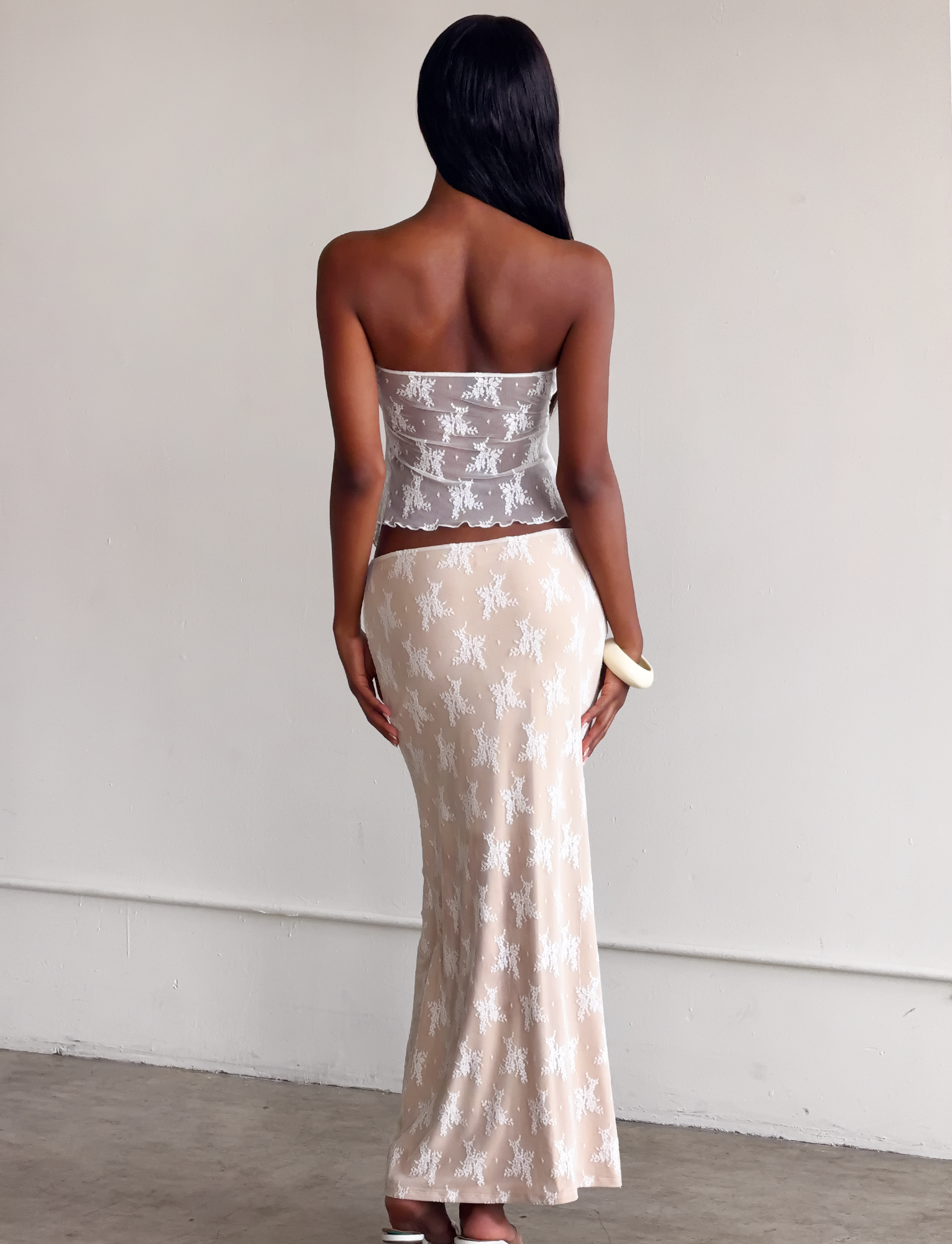 A woman with long dark hair, back to the camera, wears the TIGERMIST KAROLINA TOP in white lace and a long beige skirt, holding a small white clutch against a plain light wall.