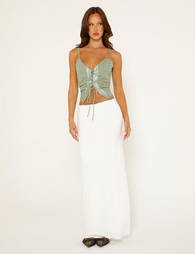 A woman stands against a plain white background, wearing a light green, ruched Tiger Mist sleeveless top with thin spaghetti straps and the TIGERMIST ADDISON SKIRT in white. Her long, wavy hair cascades over her shoulders, and she completes the look with black pointed-toe shoes.