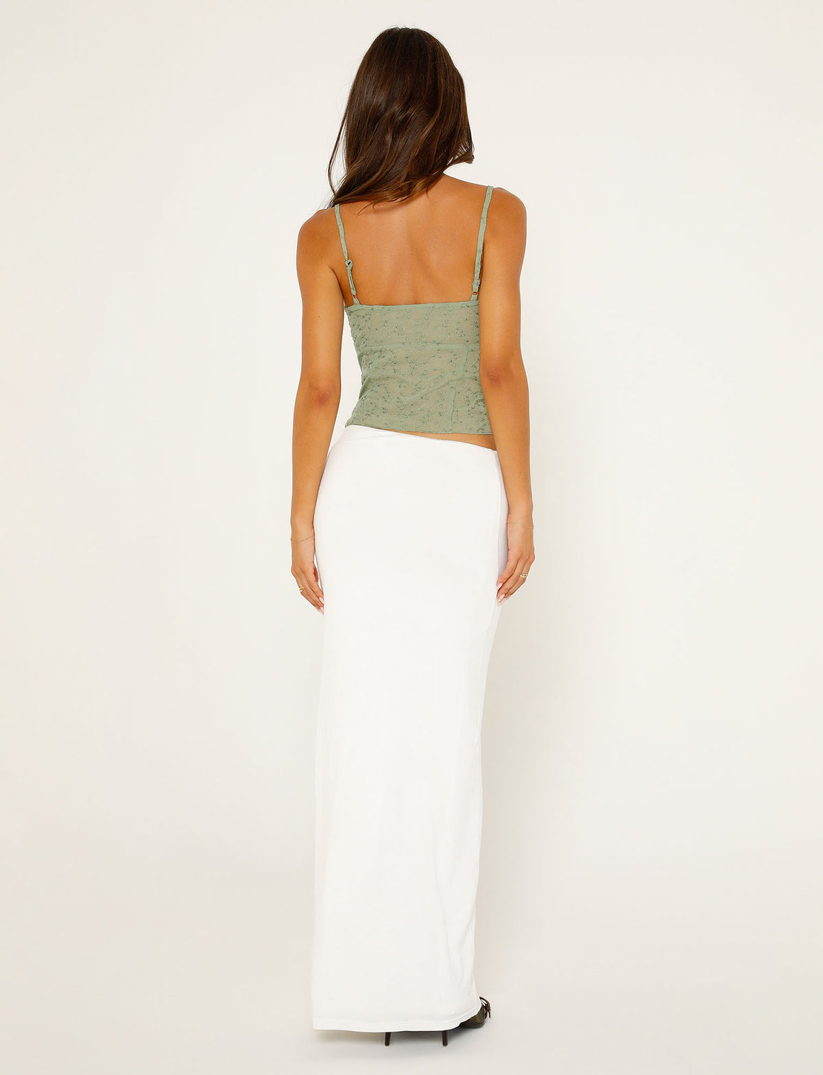 A woman with long brown hair stands, facing away from the camera. She is wearing a green spaghetti strap top and the Addison Skirt in white from TIGERMIST. The background is plain and light-colored.