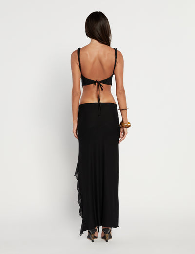 A woman with long dark hair stands facing away, wearing a striking TIGERMIST THEO TOP in black and a long black wrap skirt. She complements her look with high-heeled sandals and a wide bracelet on her left wrist. The background is plain and white.