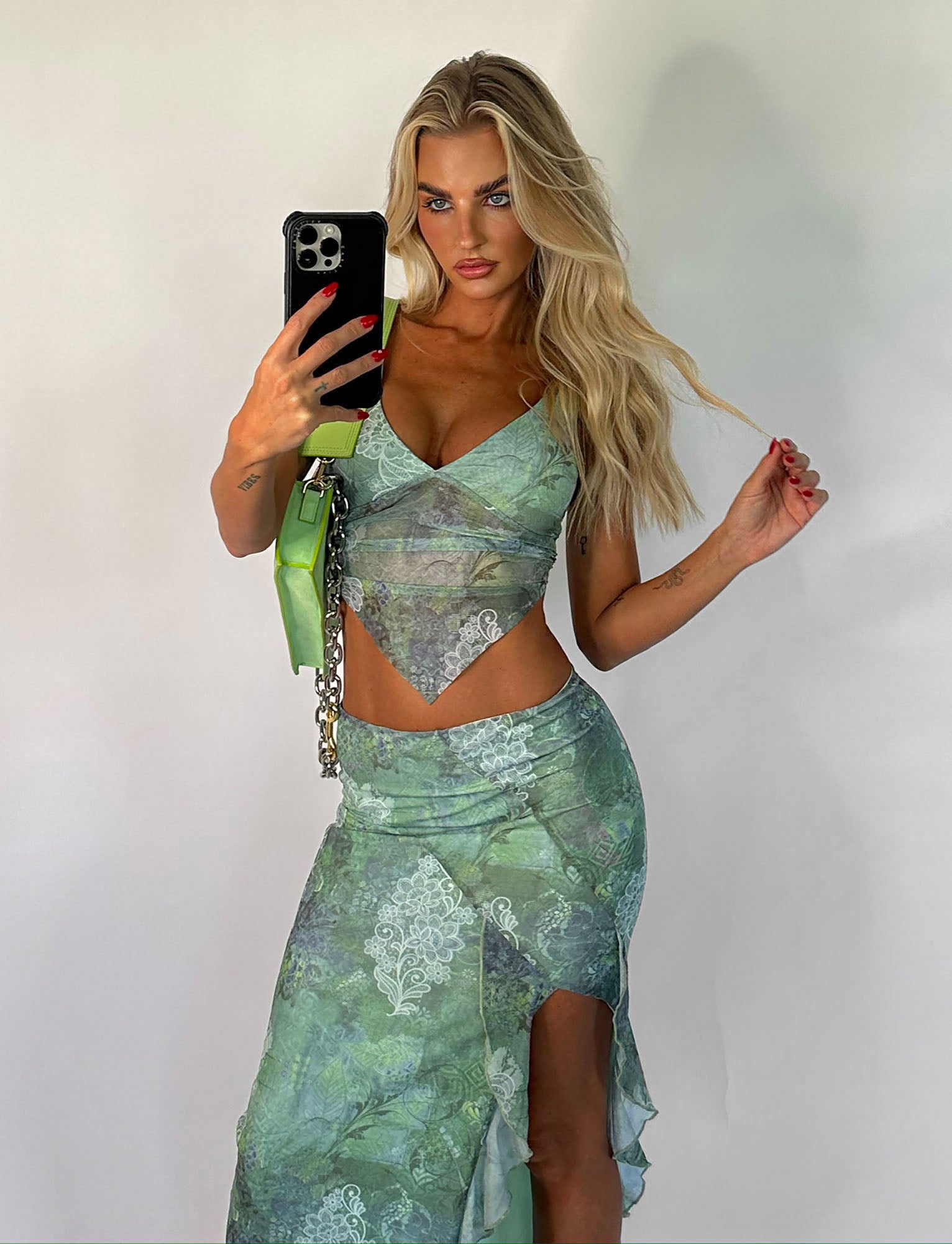 A woman with long blonde hair takes a mirror selfie, wearing a TIGERMIST outfit consisting of the THEO TOP - GREEN: FLORAL crop top and a matching skirt featuring a slit. She holds a small green purse in her right hand while her left hand touches her hair, highlighting the flattering bodycon fit of the ensemble.
