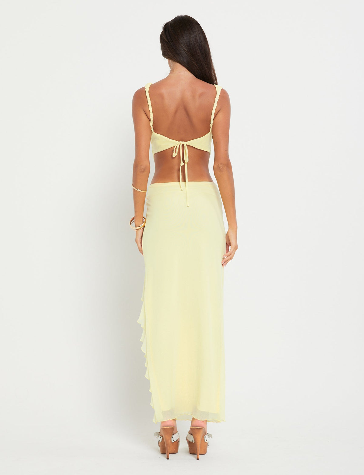 A woman with long, dark hair stands facing away. She wears a light yellow, two-piece outfit from TIGERMIST. The top features a tie-back detail, and she pairs it with the semi-sheer, long THEO SKIRT - YELLOW : LEMON. She completes her look with high-heeled sandals and minimal jewelry, including a bracelet on her right wrist.