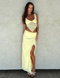 A woman stands against a light gray textured wall, wearing the light yellow LEMON two-piece outfit from TIGERMIST. Her ensemble features a cropped top and the THEO SKIRT with a high slit. She completes her look with white sandals. With long brown hair cascading down, she rests her left hand on her thigh.