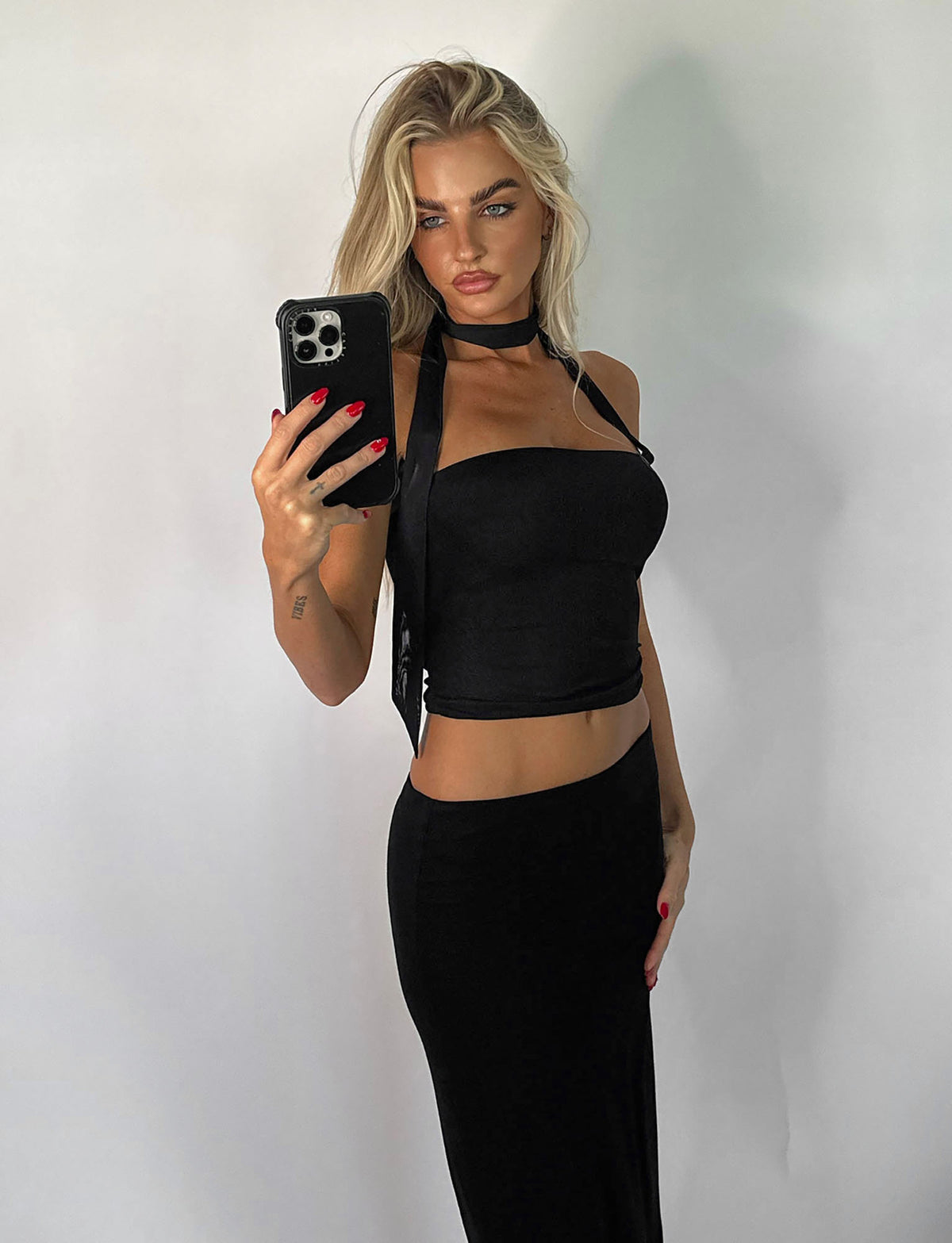 A person with long blonde hair takes a mirror selfie, rocking the CHRYSLER TOP - BLACK from TIGERMIST, paired with a black skirt and choker-style accessory. Their nails are painted red as they hold a smartphone with a black case against a plain white background.