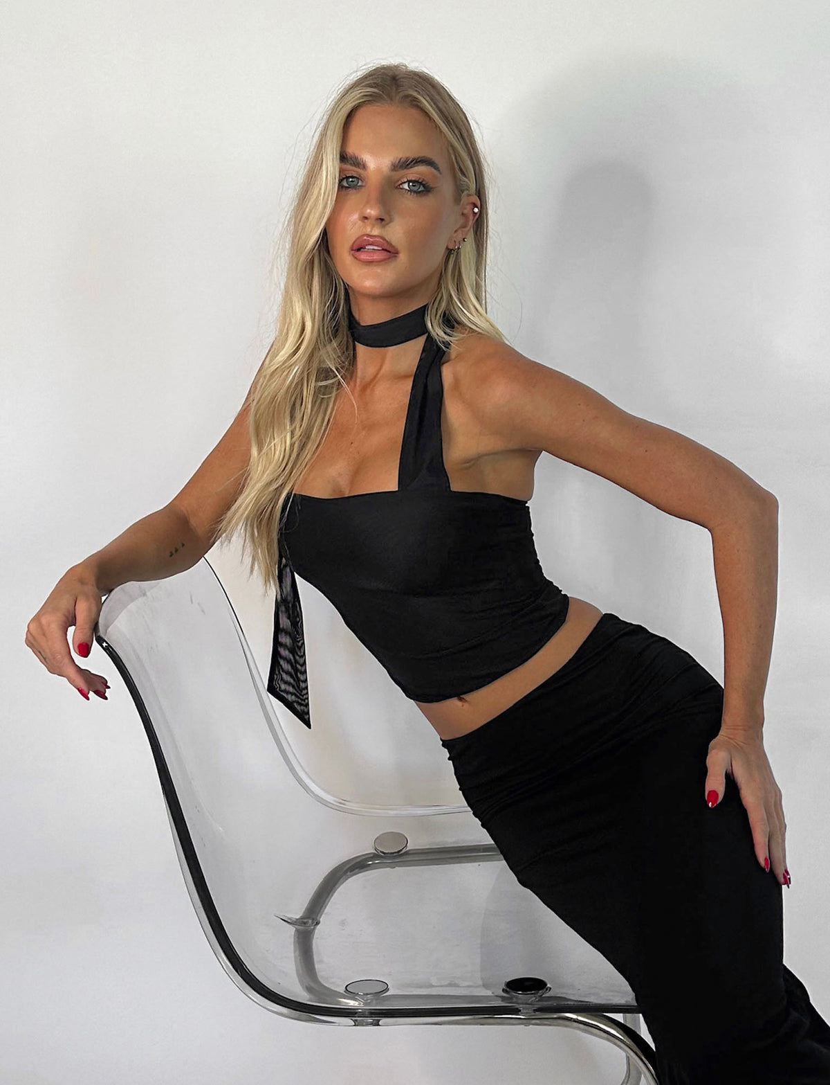 A woman with long blonde hair poses confidently in the TIGERMIST CHRYSLER TOP - BLACK and a matching black skirt. She leans against a clear plastic chair, one hand on the backrest and the other on her hip, set against a white background.