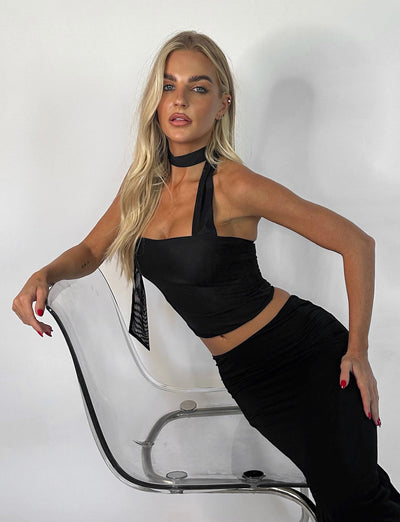 A woman with long blonde hair poses confidently in the TIGERMIST CHRYSLER TOP - BLACK and a matching black skirt. She leans against a clear plastic chair, one hand on the backrest and the other on her hip, set against a white background.