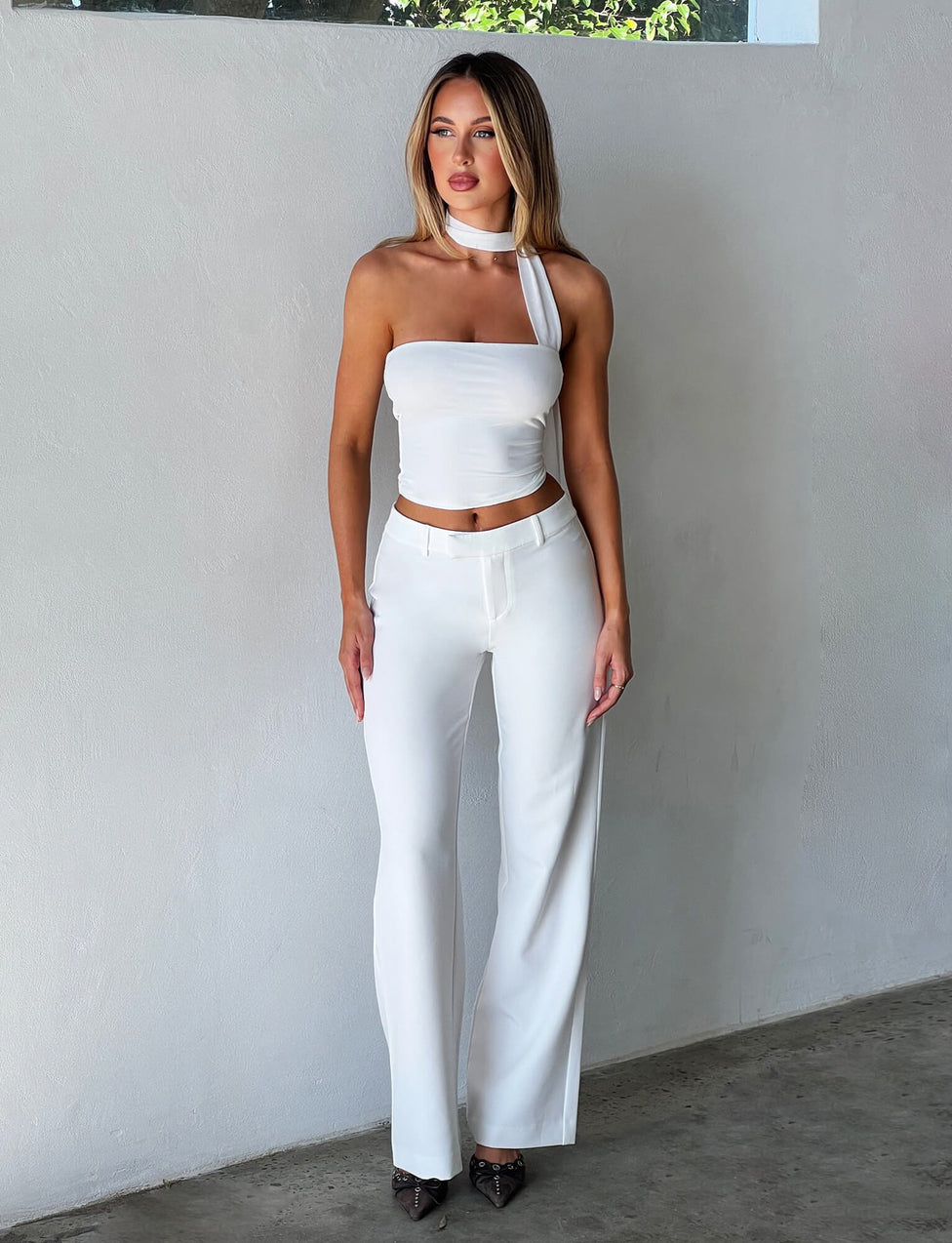 A person with long hair stands against a smooth, light-colored wall, wearing the stylish CHRYSLER TOP - WHITE from Tigermist, which features an asymmetrical neckline. They pair it with high-waisted white pants and pointed-toe dark shoes to complete the look.