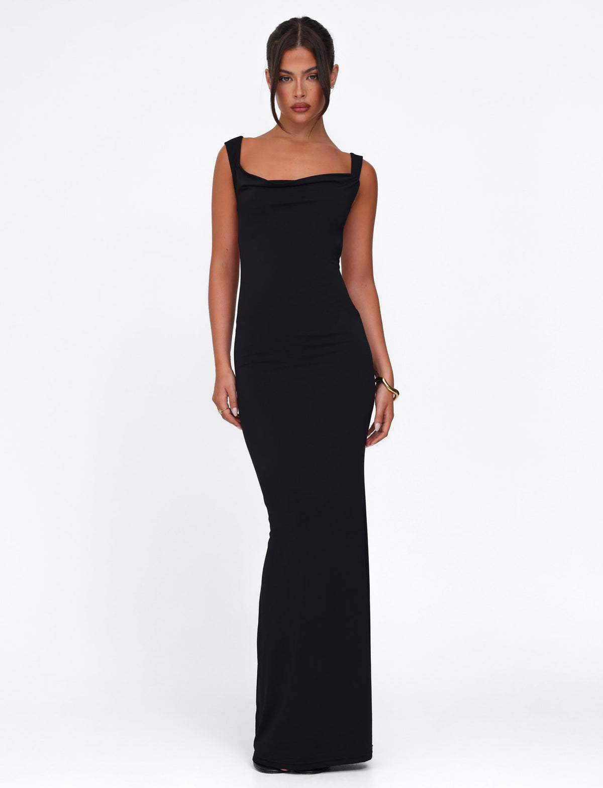 A woman stands against a white background wearing the sleek MORANA DRESS - BLACK by TIGERMIST, a long bodycon maxi dress with wide straps. Her hair is pulled back, and she displays a neutral expression.