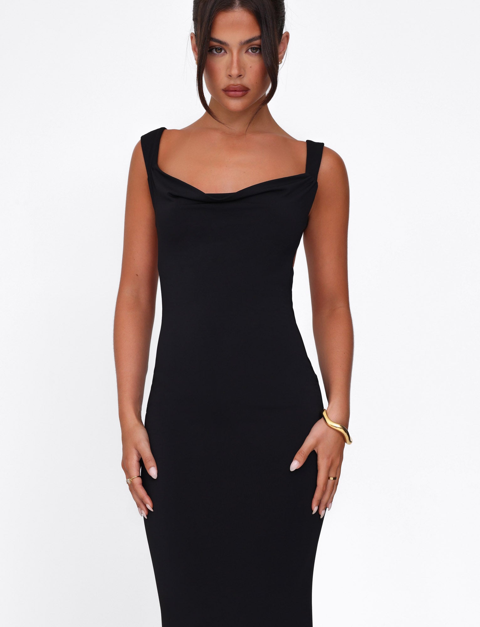 Dressed in the TIGERMIST MORANA DRESS - BLACK, she poses against a white background. Her long dark hair is styled up, and her minimal jewelry includes a bracelet and rings. With a neutral expression, her hands rest confidently on her hips.