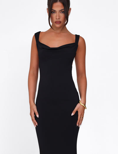 Dressed in the TIGERMIST MORANA DRESS - BLACK, she poses against a white background. Her long dark hair is styled up, and her minimal jewelry includes a bracelet and rings. With a neutral expression, her hands rest confidently on her hips.