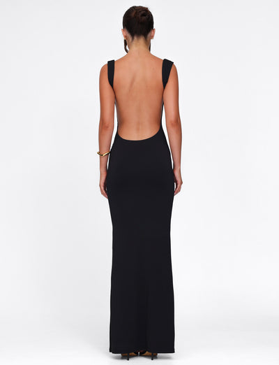 The individual gracefully wears the sleeveless, backless MORANA DRESS - BLACK by TIGERMIST, a floor-length maxi with wide straps and bodycon fit, standing elegantly against a plain white backdrop, highlighting its sophisticated silhouette as they face away from the camera.