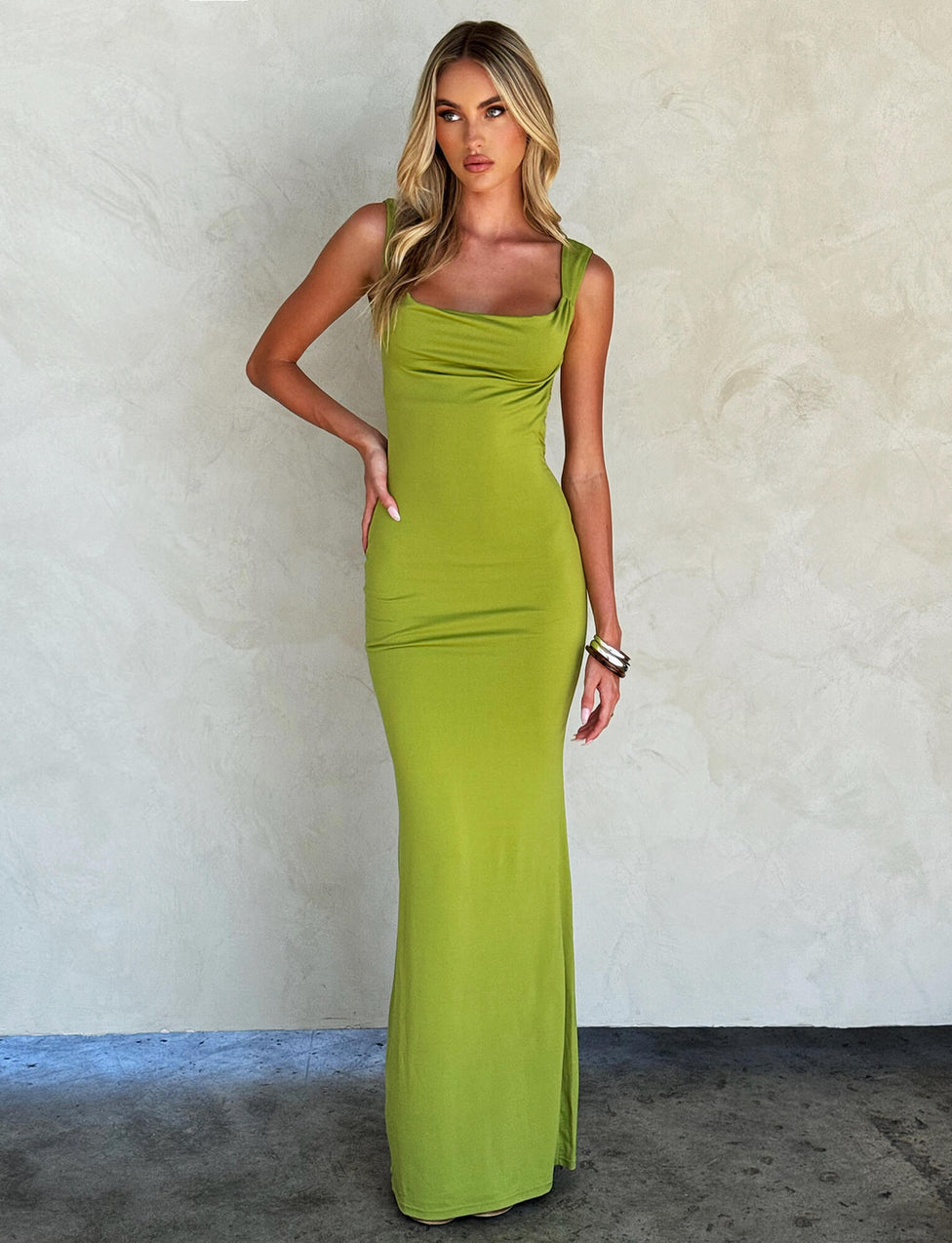 A woman with long, blonde hair is posing against a plain background. She is wearing a form-fitting, sleeveless MORANA dress in pastel green from TIGERMIST, featuring a draped neckline and a floor-length hem. She accessorizes with bangles on her right wrist.