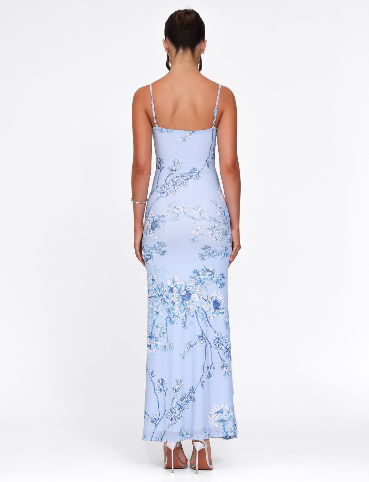 A woman faces away wearing the TIGERMIST JULIANNA DRESS in Blue Ceramic Floral—a light blue, floor-length maxi with thin straps—styled with white high-heeled sandals, her hair up, against a plain white background.