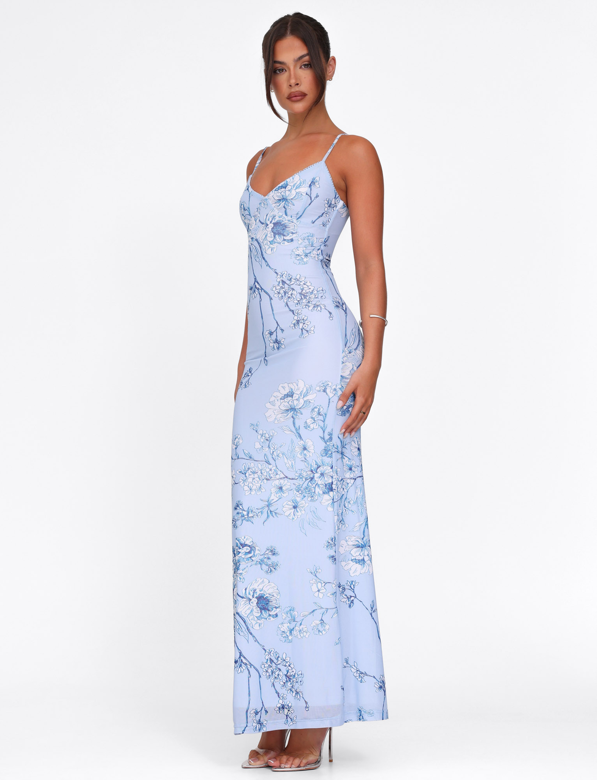 A woman with dark hair tied back wears the TIGERMIST JULIANNA DRESS in blue ceramic floral—a light blue, floor-length maxi—paired with high heels and a thin bracelet, standing against a white background.