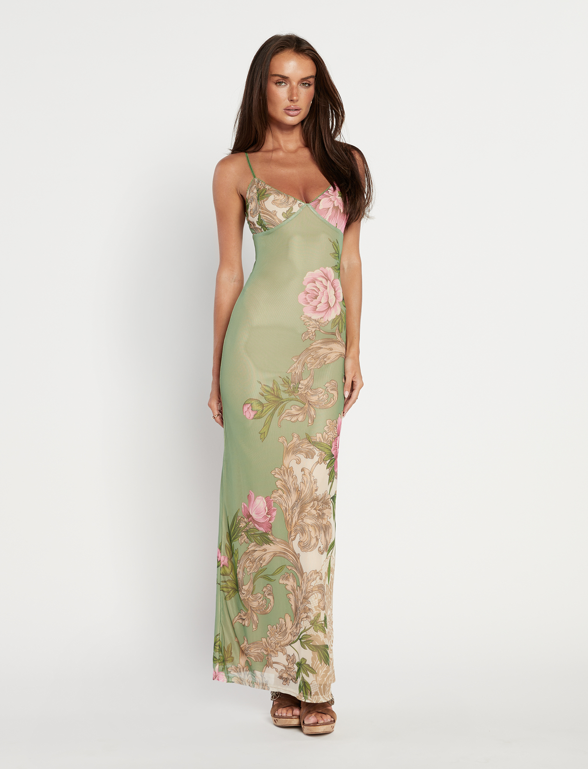A woman with long brown hair wears the TIGERMIST JULIANNA DRESS in green Rococo Floral—a sleeveless maxi with thin straps and a pink-and-beige floral pattern—paired with brown sandals, standing against a white background.