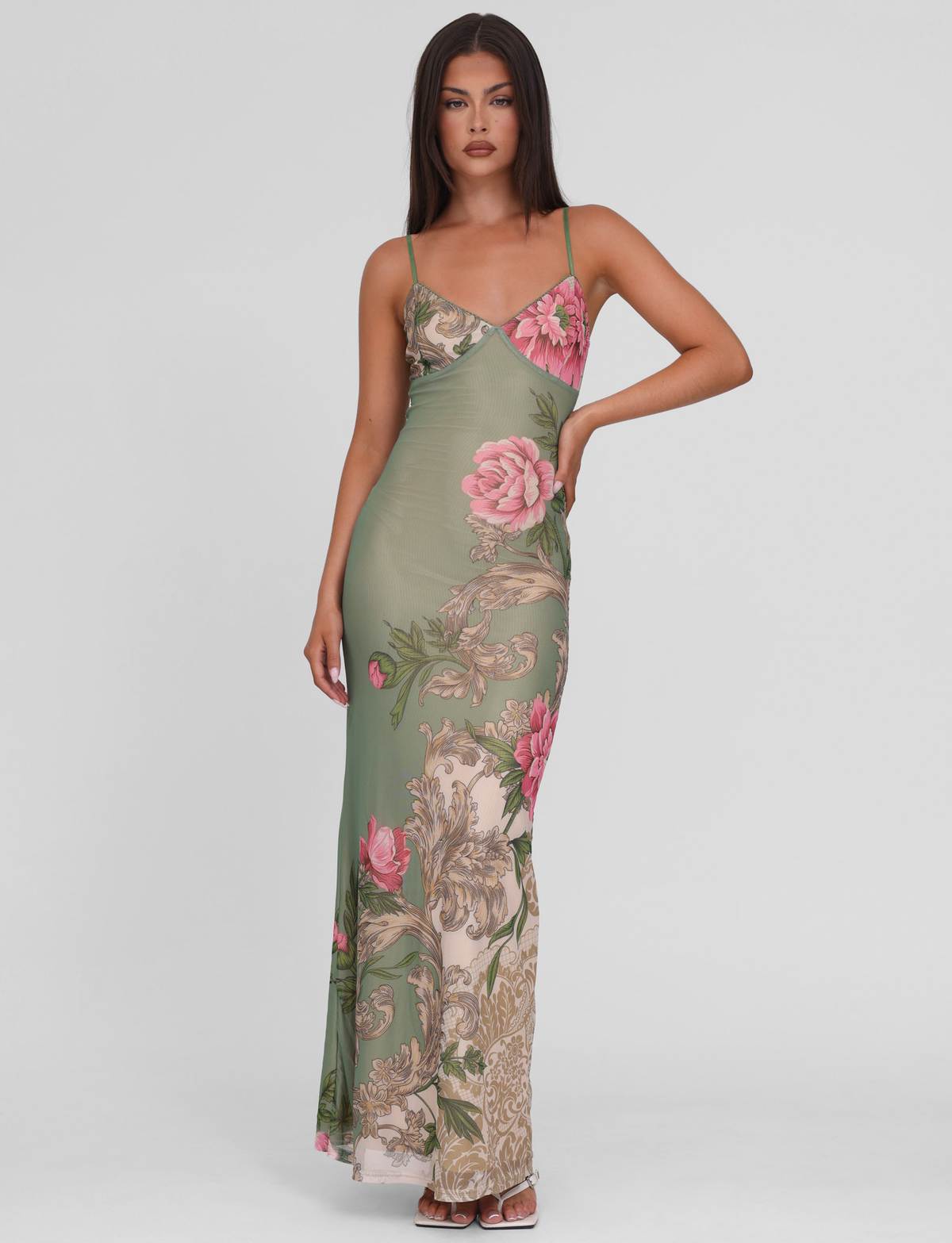 A woman with long dark hair and a neutral expression poses against a plain background, wearing the TIGERMIST JULIANNA DRESS in green Rococo floral with pink flowers and beige details; one hand rests on her hip.