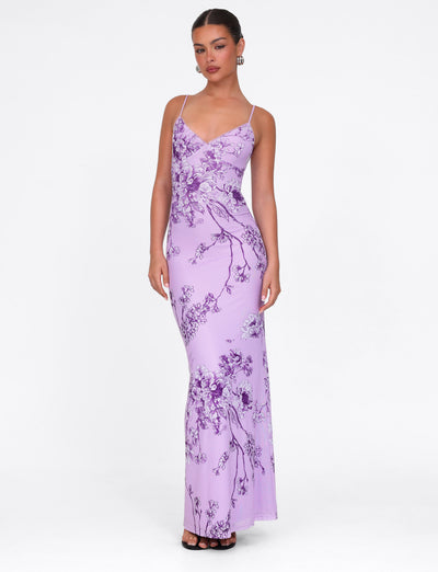 A person poses in a TIGERMIST JULIANNA DRESS - PURPLE CERAMIC FLORAL, featuring a maxi-length and thin straps, with their hair tied back and hoop earrings, against a plain white background.