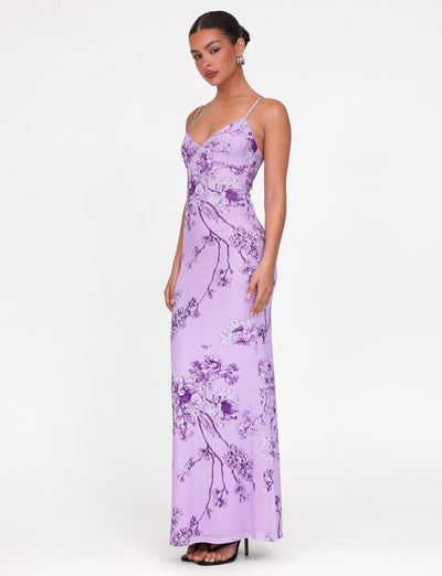Dressed elegantly in the TIGERMIST JULIANNA DRESS, a maxi-length masterpiece featuring a purple ceramic floral pattern, the woman stands against a plain backdrop. The dress's thin straps and V-neck complement her updo and black heels perfectly.