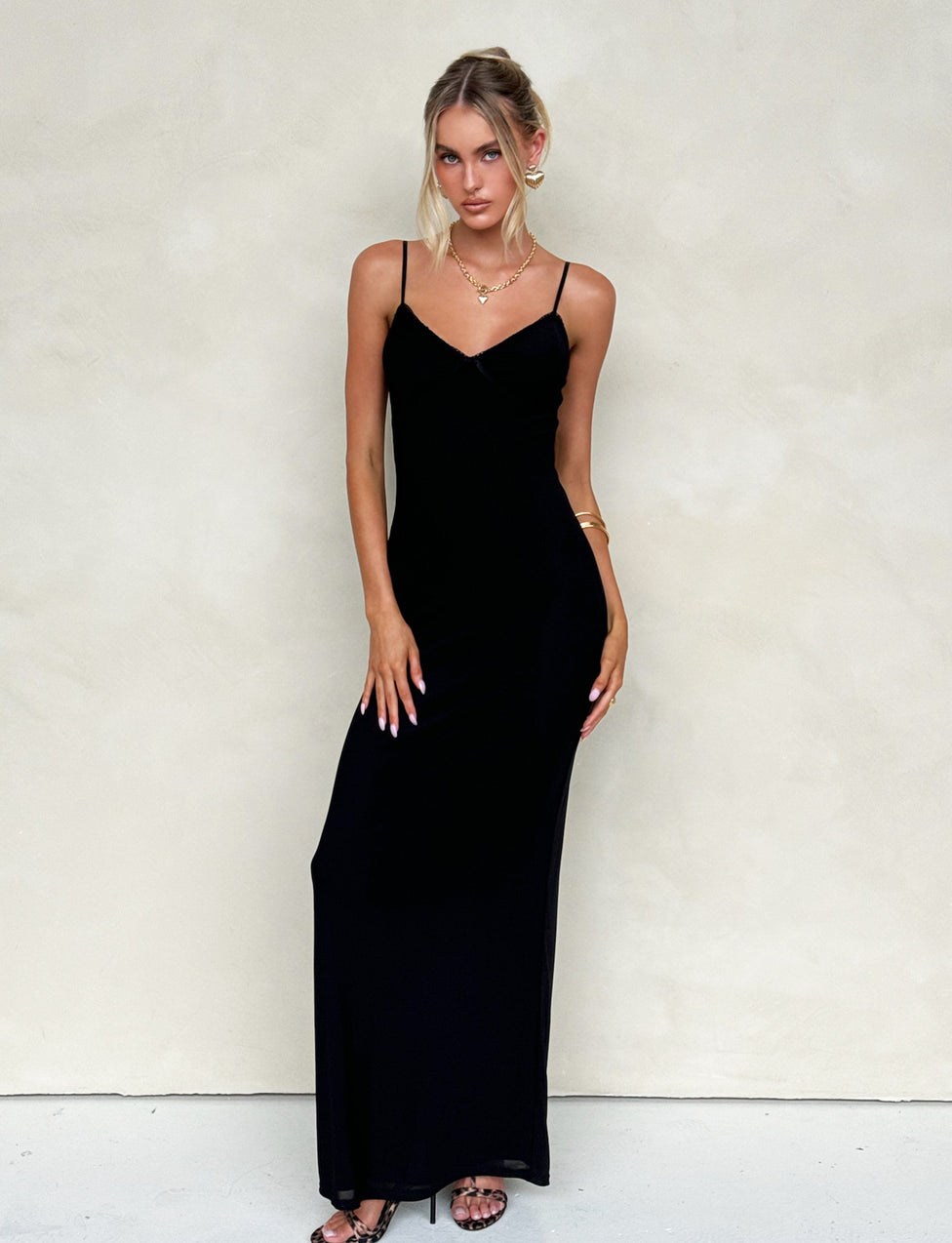A woman stands against a plain beige background, wearing the TIGERMIST JULIANNA DRESS - BLACK, a long black spaghetti-strap maxi dress with subtle eyelet trim. She has blonde hair styled in a loose updo and accessorizes with gold jewelry, including a necklace and bracelet. She poses with one hand on her hip and the other hanging by her side.