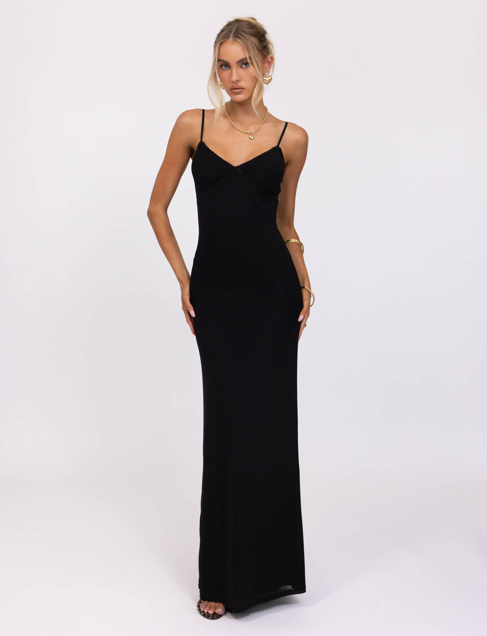 A woman stands against a plain white background, wearing the JULIANNA DRESS - BLACK by TIGERMIST with thin straps. Her hair is styled in a loose updo, and she accessorizes with a necklace, bracelet, and rings. She holds one hand by her side while the other gently touches her hip.