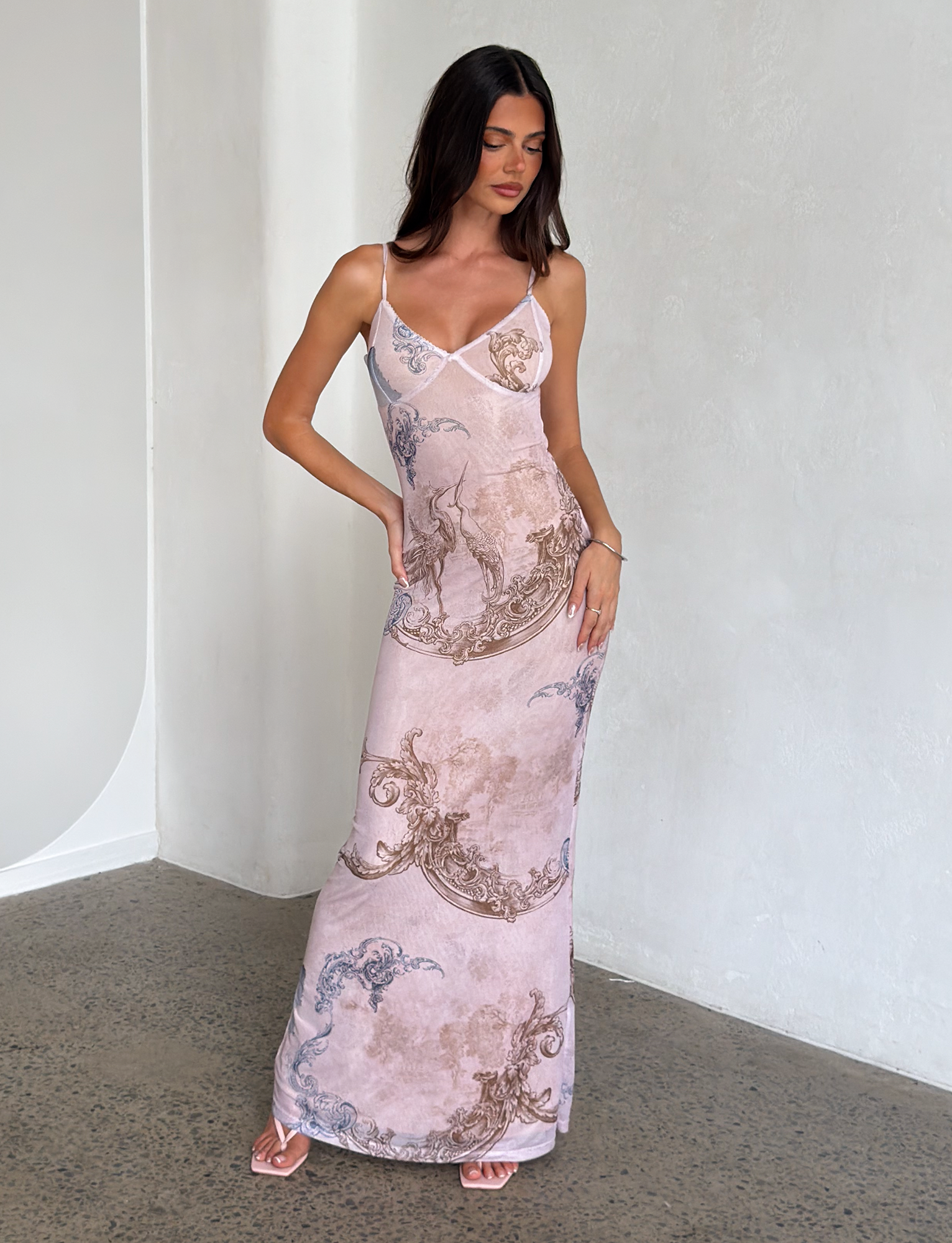 A woman with long dark hair wears the TIGERMIST JULIANNA DRESS - MULTI: COLLAGE: CRANES, a sleeveless printed maxi dress with crane and collage patterns. She stands indoors against a light-textured wall, looking down with one hand on her hip.