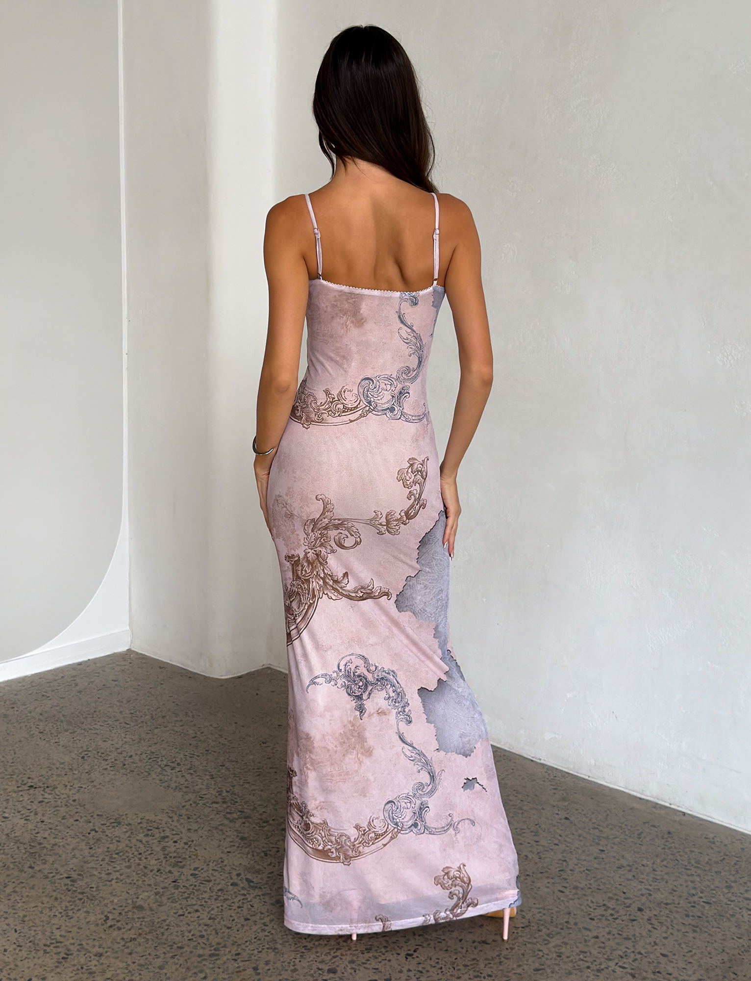 A woman with long dark hair stands indoors facing a white wall, seen from the back, wearing the TIGERMIST JULIANNA DRESS - MULTI: COLLAGE: CRANES—a fitted, sleeveless printed maxi dress.