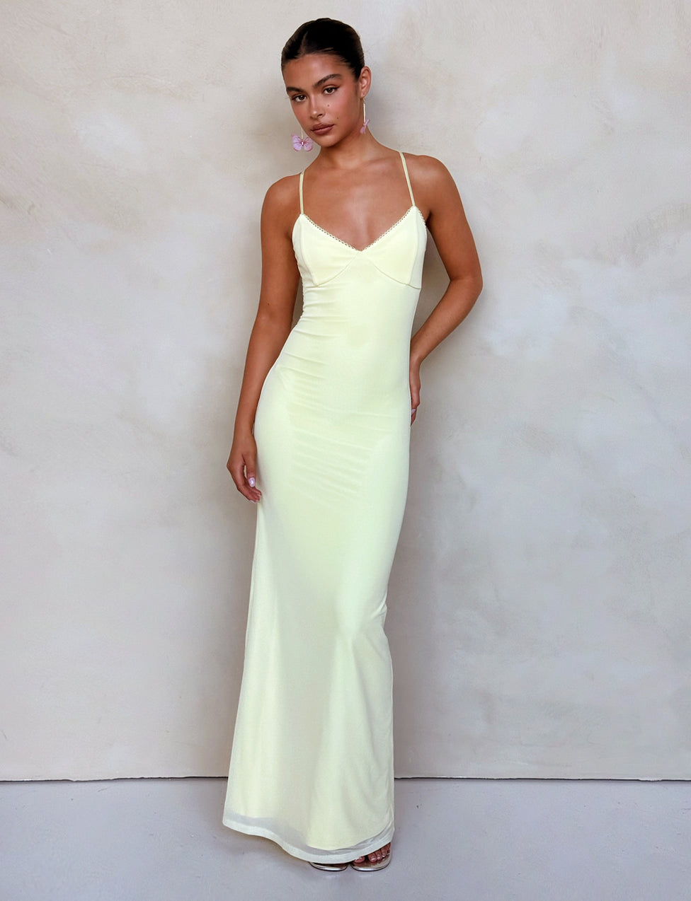 A woman stands against a plain backdrop wearing the Tiger Mist JULIANNA DRESS in lemon, a long, pale yellow, form-fitting dress with spaghetti straps. She has her hair pulled back and is looking toward the camera. One arm rests at her side while the other is slightly raised, holding the dress.