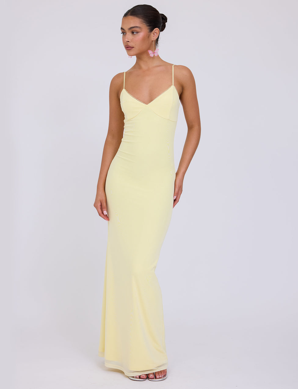 A woman is wearing the JULIANNA DRESS - YELLOW : LEMON from TIGERMIST. She has her hair styled in a sleek bun and is accessorized with dangling pink earrings. She stands against a plain white background, looking slightly to her left.