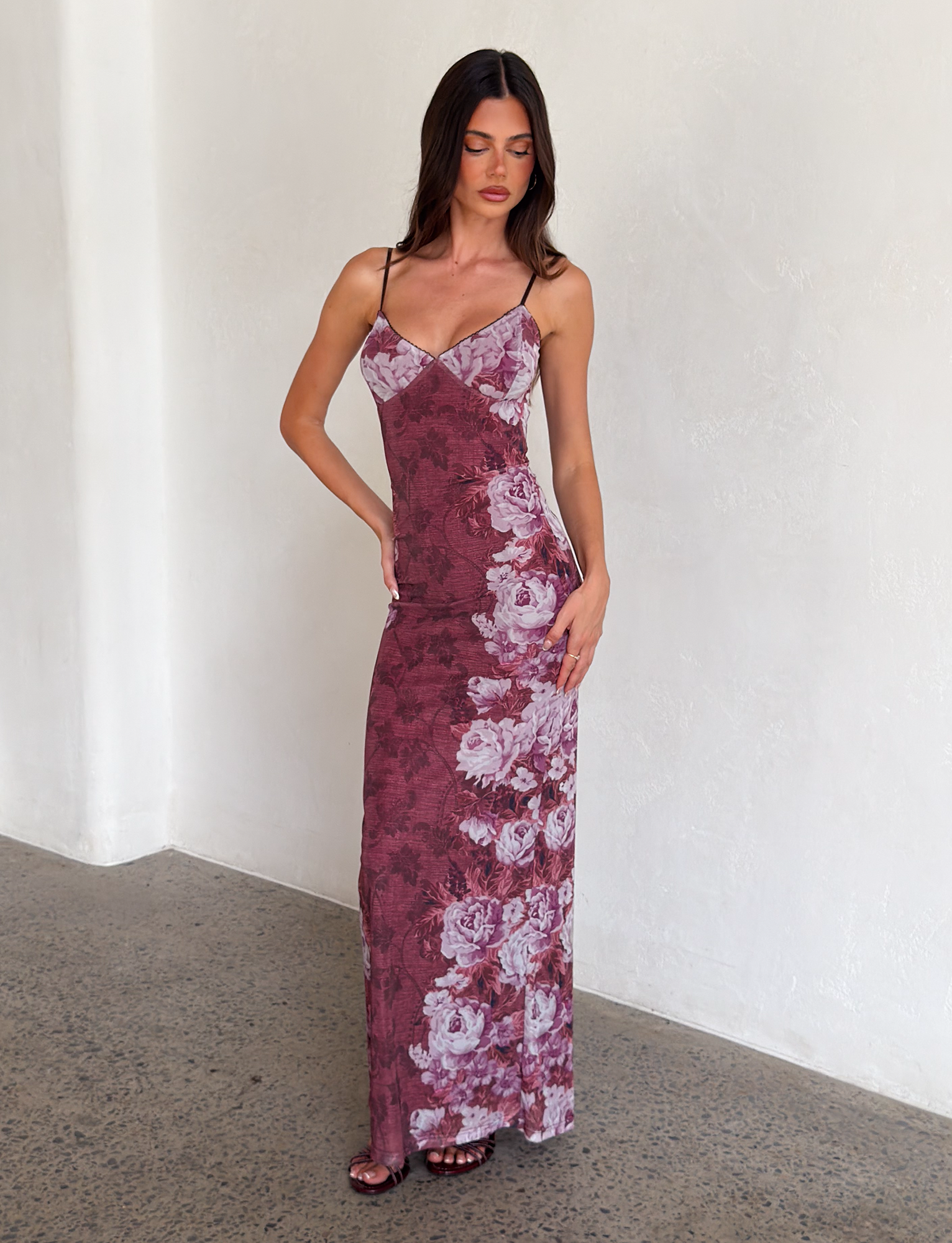 A woman stands indoors against a light wall, wearing the JULIANNA DRESS - MULTI: FLORAL: ROSE STRIPE by TIGERMIST—a long, fitted dress with thin straps and a purple floral stripe print. She looks down with one hand on her hip.