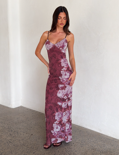 A woman stands indoors against a light wall, wearing the JULIANNA DRESS - MULTI: FLORAL: ROSE STRIPE by TIGERMIST—a long, fitted dress with thin straps and a purple floral stripe print. She looks down with one hand on her hip.