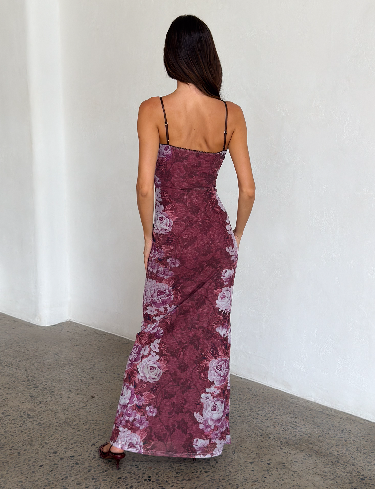 A woman with long brown hair faces away, wearing TIGERMIST's JULIANNA DRESS in Multi Floral Rose Stripe, featuring thin straps, against a white textured wall and stone floor.