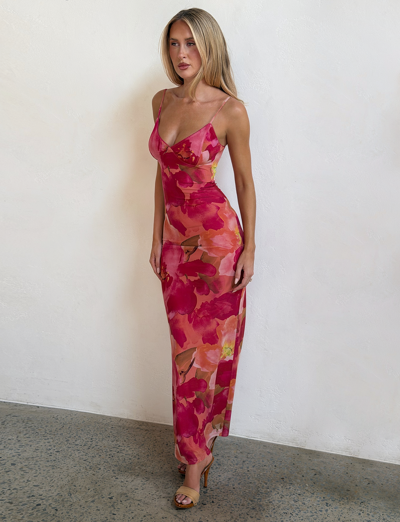A woman stands on a concrete floor against a white textured wall, wearing the Tiger Mist CL - JULIANNA DRESS, a fitted, sleeveless, ankle-length dress with a pink and red floral pattern, paired with strappy heels.