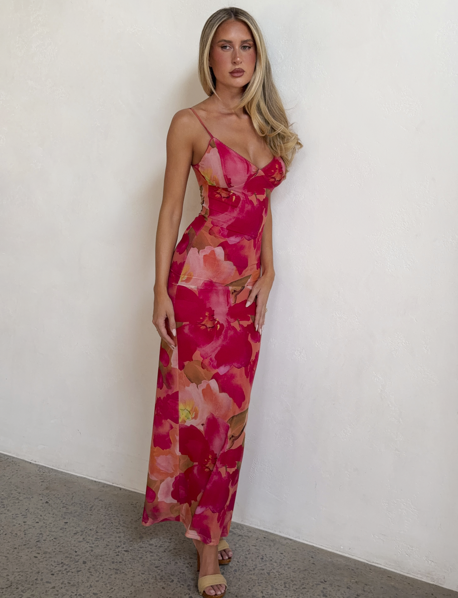 A woman with long blonde hair wears the TIGERMIST JULIANNA MAXI DRESS - DAIQUIRI FLORAL, featuring a red and pink floral print and adjustable spaghetti straps, paired with beige open-toe heels, standing against a white wall on a concrete floor.