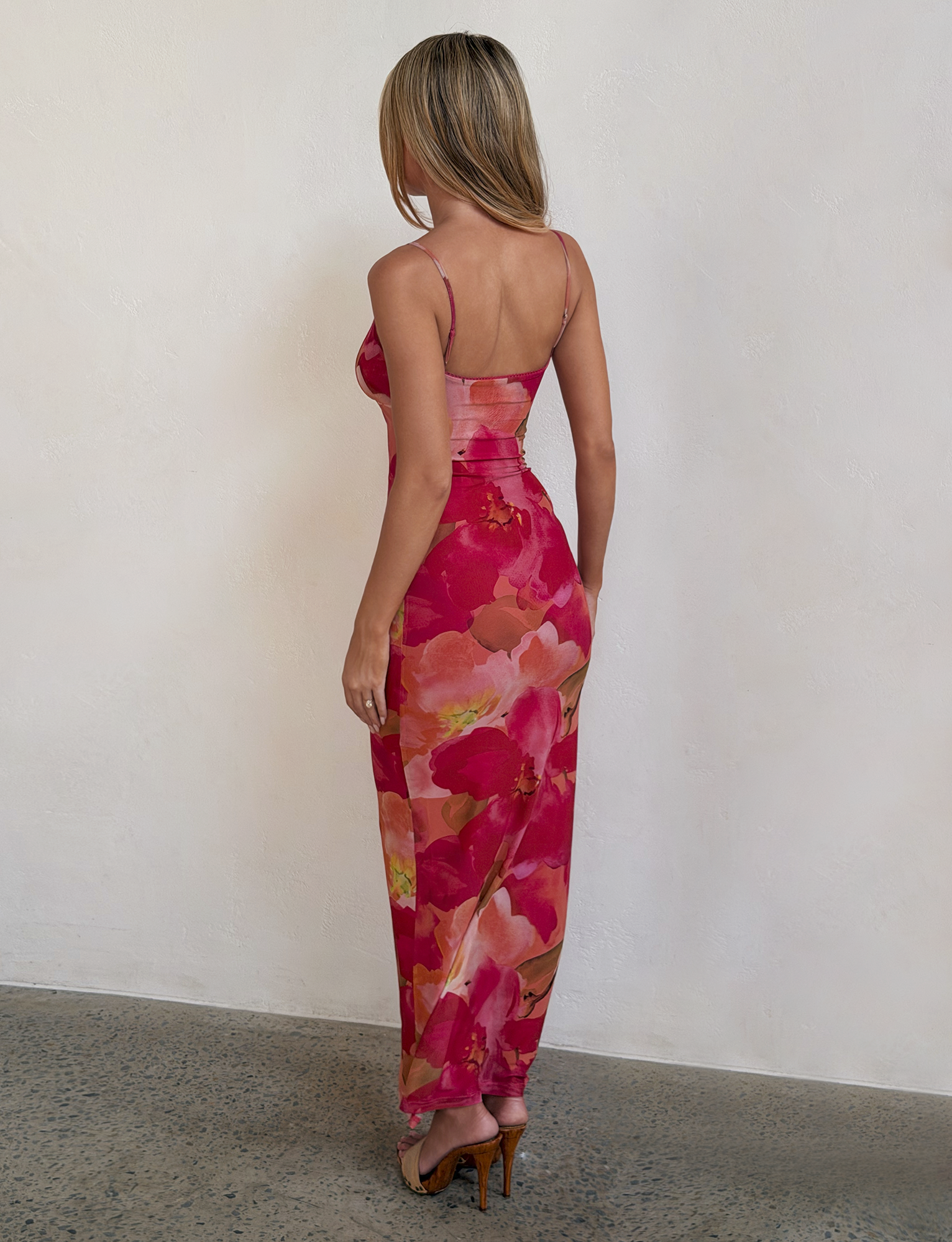 A woman with long blonde hair stands indoors, facing away, wearing the TIGERMIST JULIANNA MAXI DRESS - DAIQUIRI FLORAL with adjustable spaghetti straps and brown heels, set against a plain white wall and speckled floor.