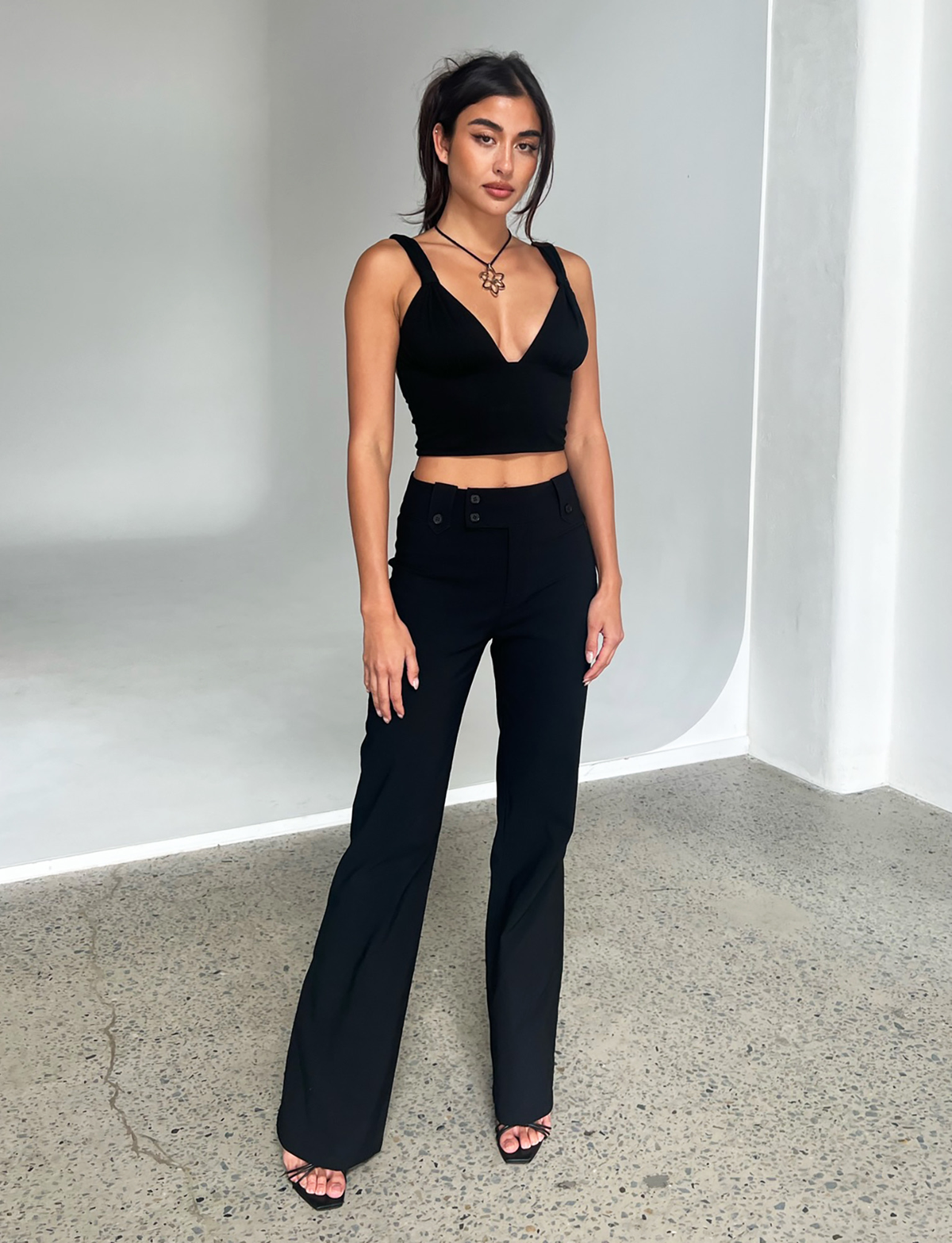 A woman stands indoors on a concrete floor wearing TIGERMIST’s KITTIE HIGH RISE PANT in black, styled with a black sleeveless crop top and heels. She has long dark hair tied back, wears a necklace, and poses against a minimal white background.
