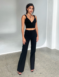A woman stands indoors on a concrete floor wearing TIGERMIST’s KITTIE HIGH RISE PANT in black, styled with a black sleeveless crop top and heels. She has long dark hair tied back, wears a necklace, and poses against a minimal white background.
