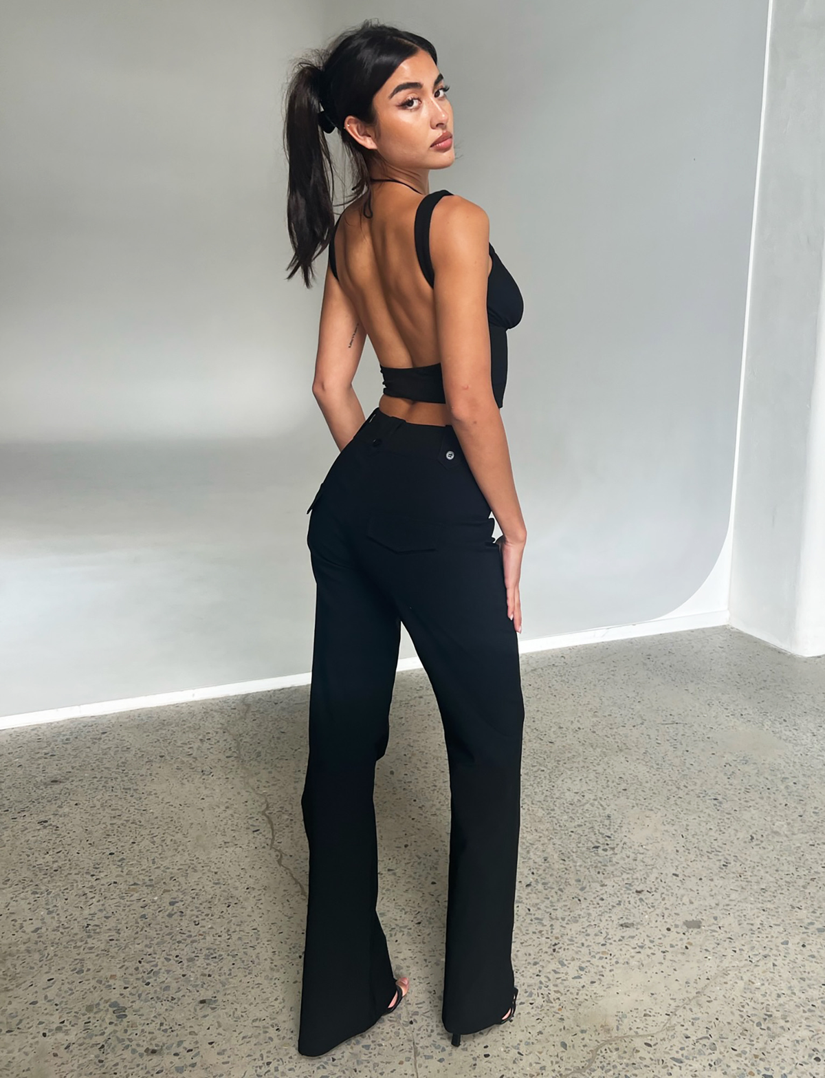 A woman with long dark hair in a ponytail poses indoors, looking over her shoulder. She wears TIGERMIST's KITTIE HIGH RISE PANT - BLACK and a black backless top, standing on a speckled concrete floor.