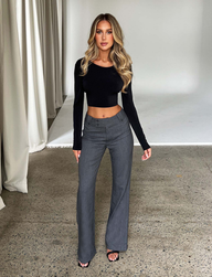 A woman with long, wavy blonde hair poses indoors in a fitted black long-sleeve crop top and TIGERMIST KITTIE HIGH RISE PANT - GREY with a flared leg, styled with open-toe heels against a neutral curtain and white wall backdrop.