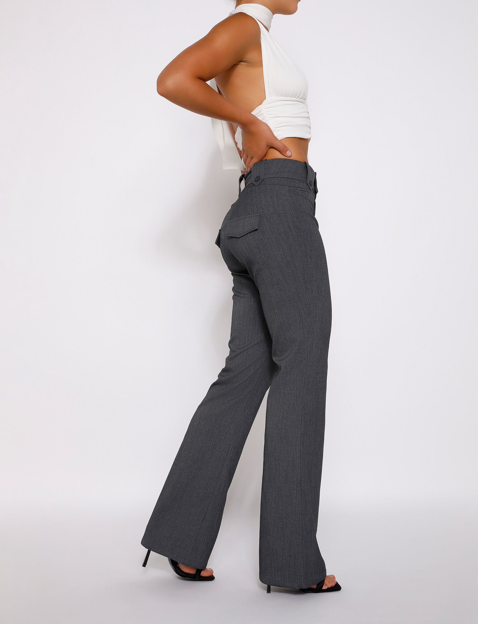 A person poses sideways against a white background, wearing the TIGERMIST KITTIE HIGH RISE PANT - GREY with a flared silhouette, paired with a sleeveless white open-back crop top and black high-heeled sandals.