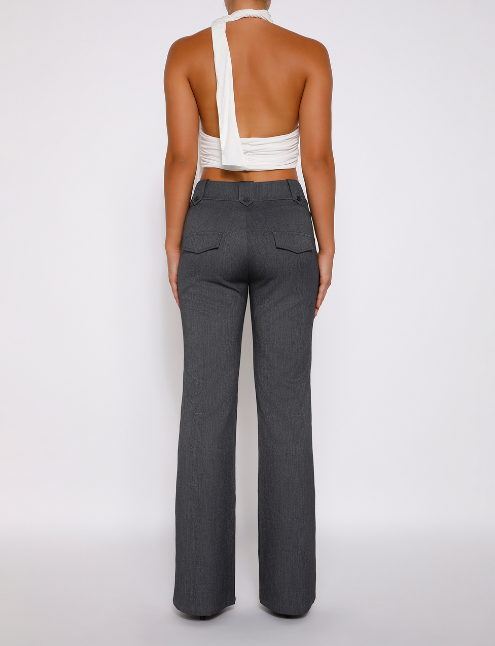 A person stands facing away against a plain white background, wearing TIGERMIST's KITTIE HIGH RISE PANT - GREY with an asymmetrical waistband and back pockets, paired with a white asymmetrical halter top.