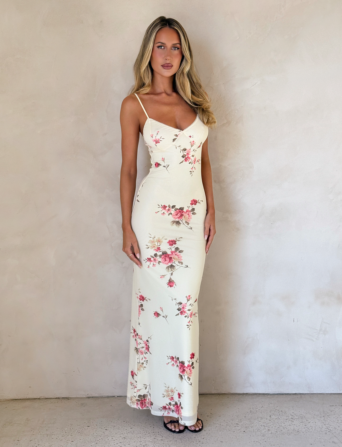 A woman wears the TIGERMIST JULIANNA DRESS in yellow macro floral print, standing against a light textured wall. Her long wavy blonde hair flows down, and she pairs the look with black strappy sandals.