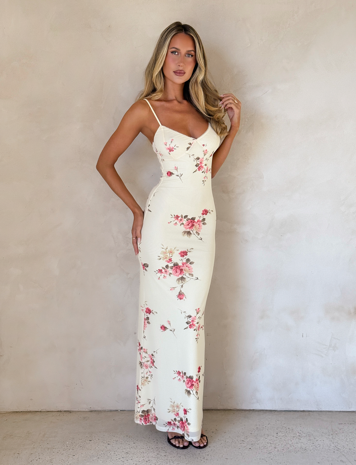A woman with long blonde hair poses by a beige wall, wearing the TIGERMIST JULIANNA DRESS in yellow macro pop floral print and black sandals, with one hand near her face.